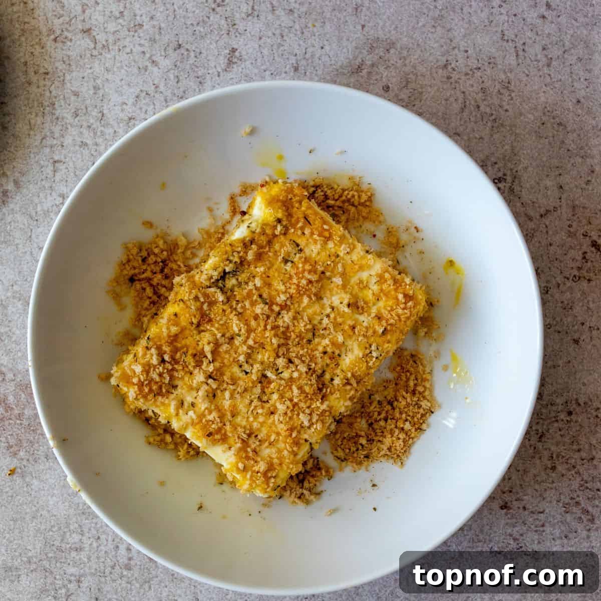 Golden Feta with Fiery Honey Drizzle 4 A block of feta cheese meticulously coated in a seasoned panko breadcrumb mixture, ready for pan frying, resting in a bowl.