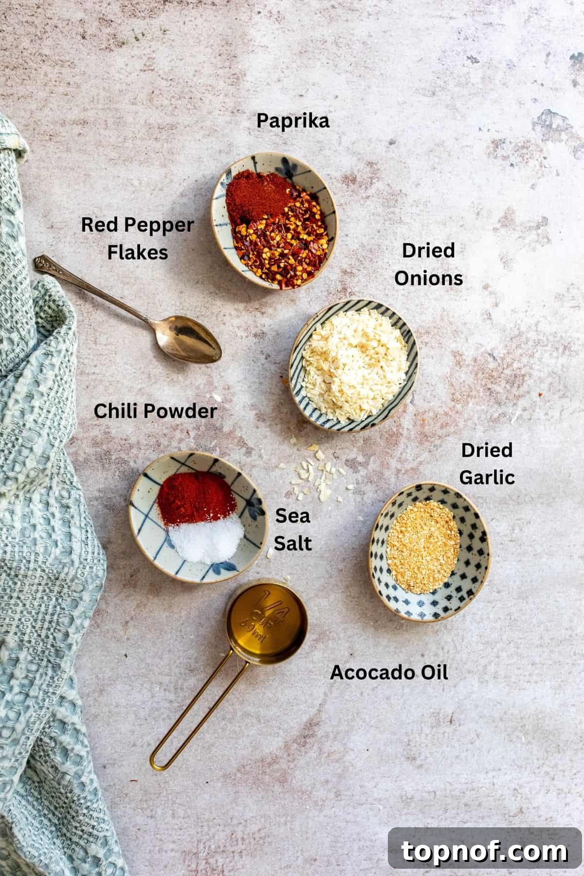 Homemade Trader Joe's Chili Onion Crunch 3 Various ingredients for Chili Onion Crunch, including dried minced garlic, dried onion, chili flakes, paprika, salt, chili powder, and a bottle of olive oil, neatly arranged in small white bowls on a light-colored kitchen counter.