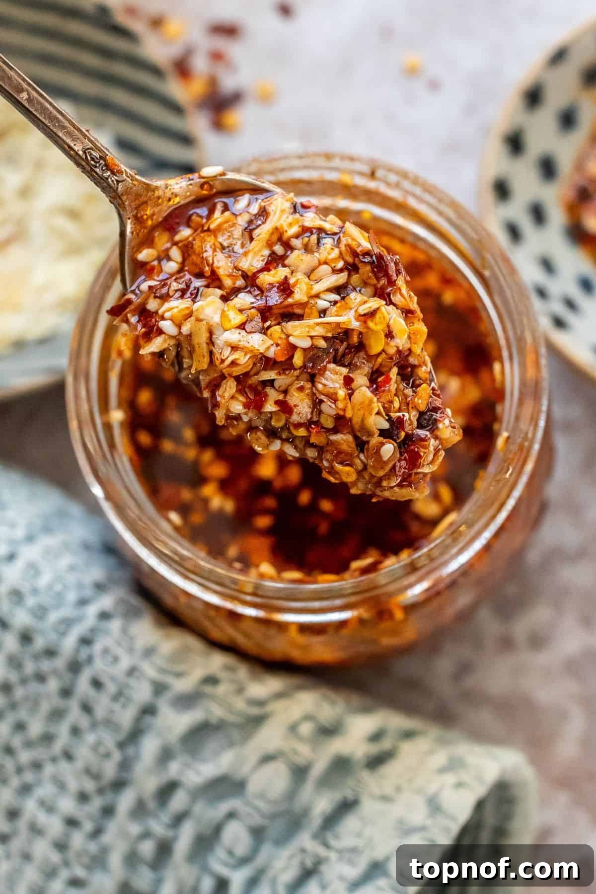 Homemade Trader Joe's Chili Onion Crunch 5 A close-up shot of a silver spoon lifting a generous amount of Chili Onion Crunch from a clear glass jar, with the texture and oil glistening invitingly.