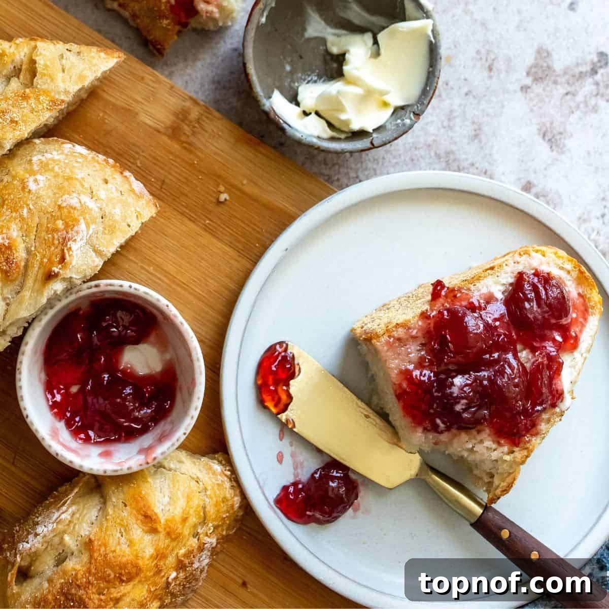 Artisan Overnight Loaf 6 A slice of golden, crusty overnight bread with a dollop of strawberry jam, placed on a white plate next to the main loaf.