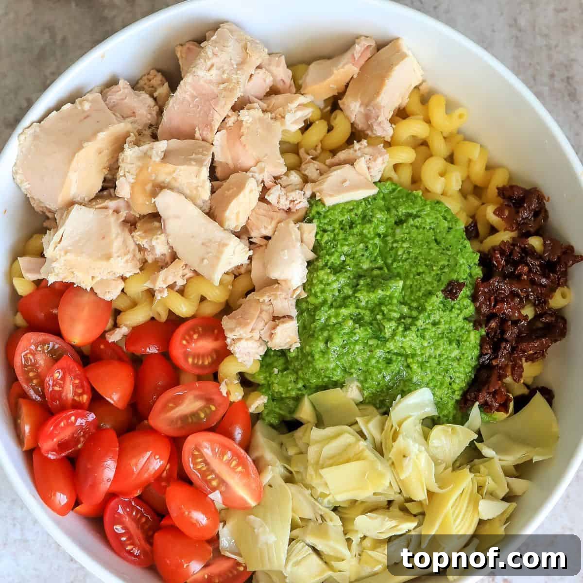 Mediterranean Tuna Pesto Pasta 5 A large white ceramic mixing bowl filled with the newly combined Tuna Pesto Pasta. Cooked pasta is generously coated in green pesto, intertwined with flaky tuna, bright cherry tomatoes, and tender artichoke hearts, ready for a final toss.