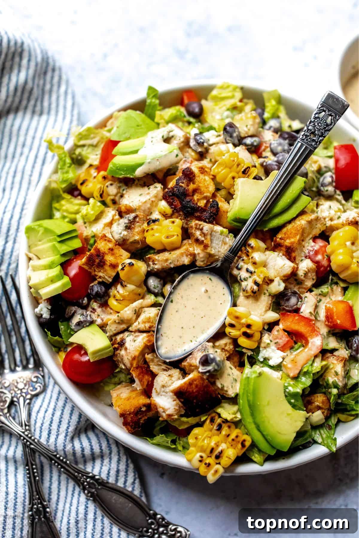 A beautifully arranged Spicy Southwest Salad in a pristine white bowl, resting on a rustic counter with a folded kitchen towel nearby. A spoonful of the creamy dressing is poised to be drizzled over, highlighting its rich texture.