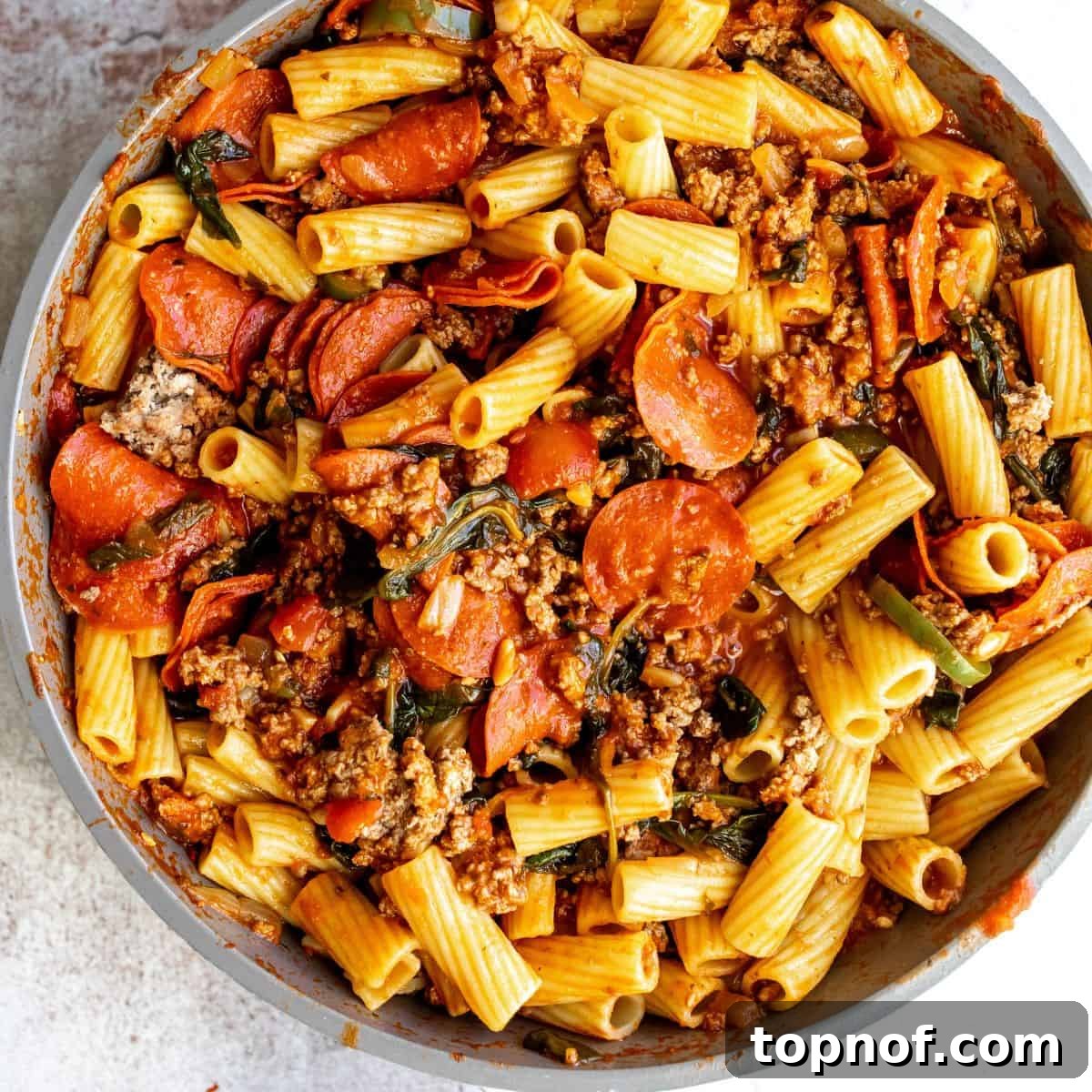 A large grey skillet filled with bubbling pepperoni pasta and a rich red sauce, featuring visible pepperoni slices and ground meat, simmering on a stovetop.