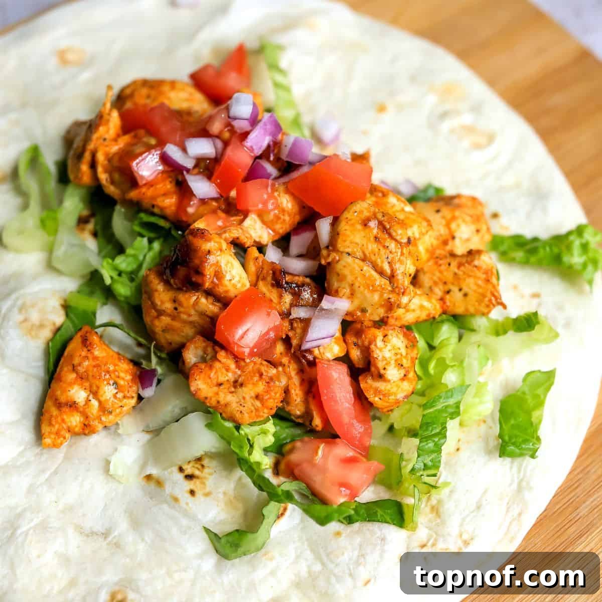 A large flour tortilla laid flat, filled with a generous portion of cooked Cajun chicken, fresh shredded lettuce, diced tomatoes, and red onions, ready for the sauce and folding.
