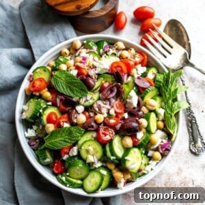 Greek cucumber salad in a white bowl on a table with silver serving utensils to the side.