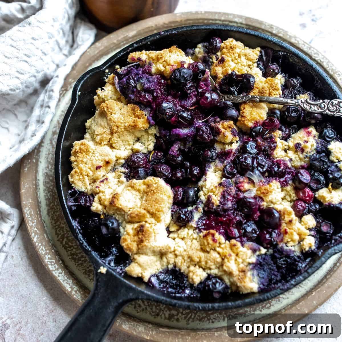 Luscious Gluten-Free Blueberry Cobbler 6 A black skillet containing the baked gluten-free blueberry cobbler, with a silver spoon scooping out a portion.