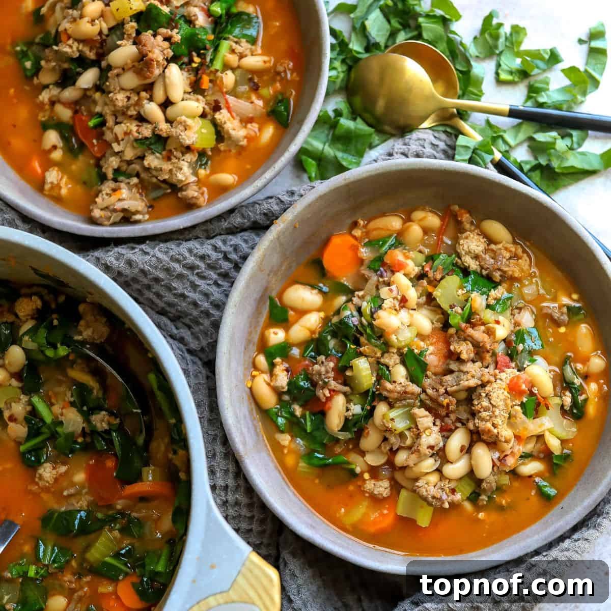 Rustic Tuscan White Bean Stew 2 A rustic grey ceramic bowl filled with hearty Tuscan white bean soup, featuring visible ground meat, white beans, and vibrant greens, with a spoon resting in the bowl, inviting you to enjoy.