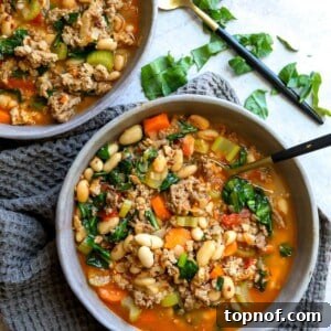 A cozy grey bowl filled with hearty Tuscan white bean soup, showing ground meat, beans, and fresh greens, accompanied by a black spoon, ready to be enjoyed.