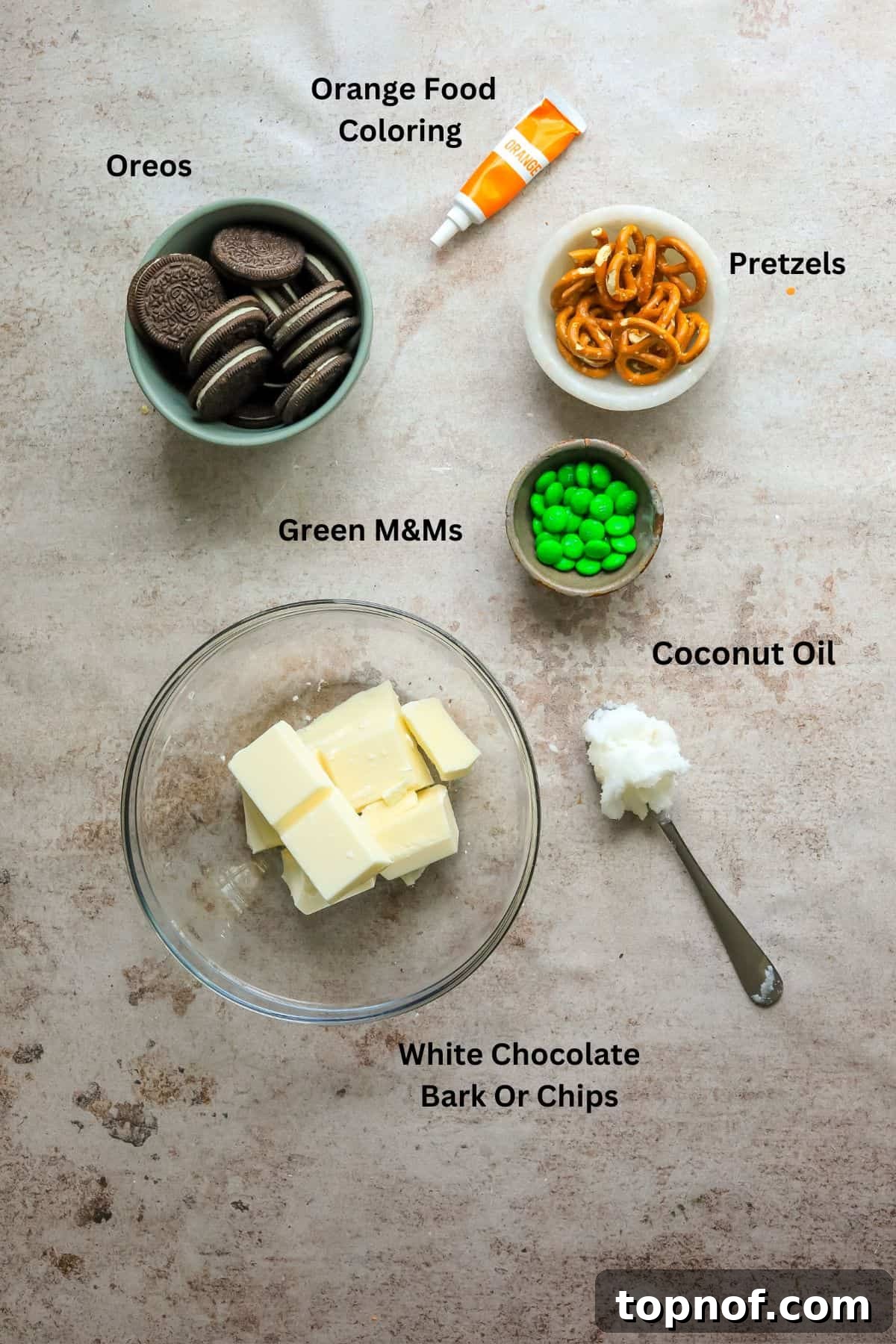 A collection of ingredients laid out, including a package of Oreo cookies, white chocolate bark, a jar of coconut oil, orange food coloring, green M&Ms, and pretzel sticks, ready for no-bake cookie preparation.
