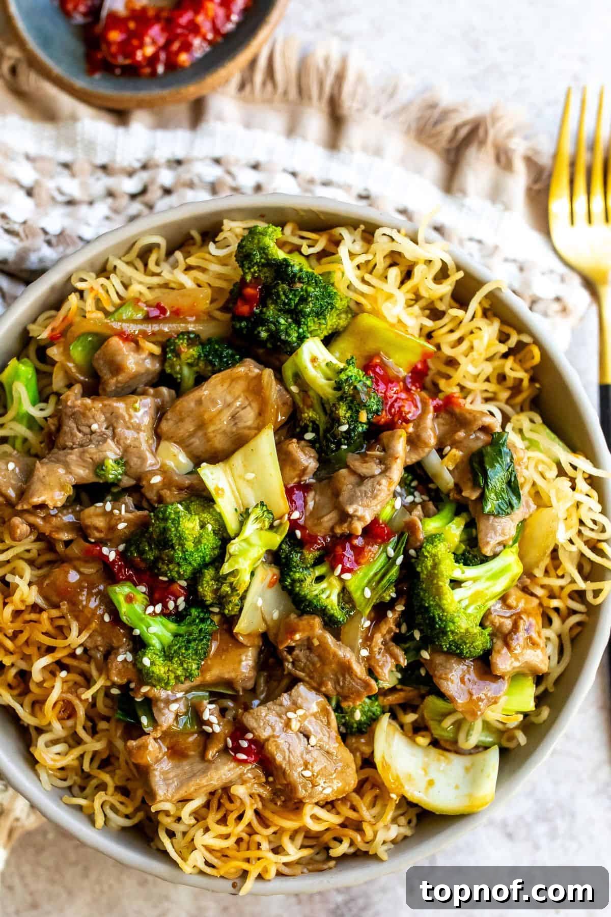 Sizzling Beef and Broccoli Noodles 2 A white bowl brimming with golden-brown crispy noodles, succulent beef strips, and vibrant green broccoli florets, coated in a rich, glossy red sauce. A fork is tucked into the side of the noodles.