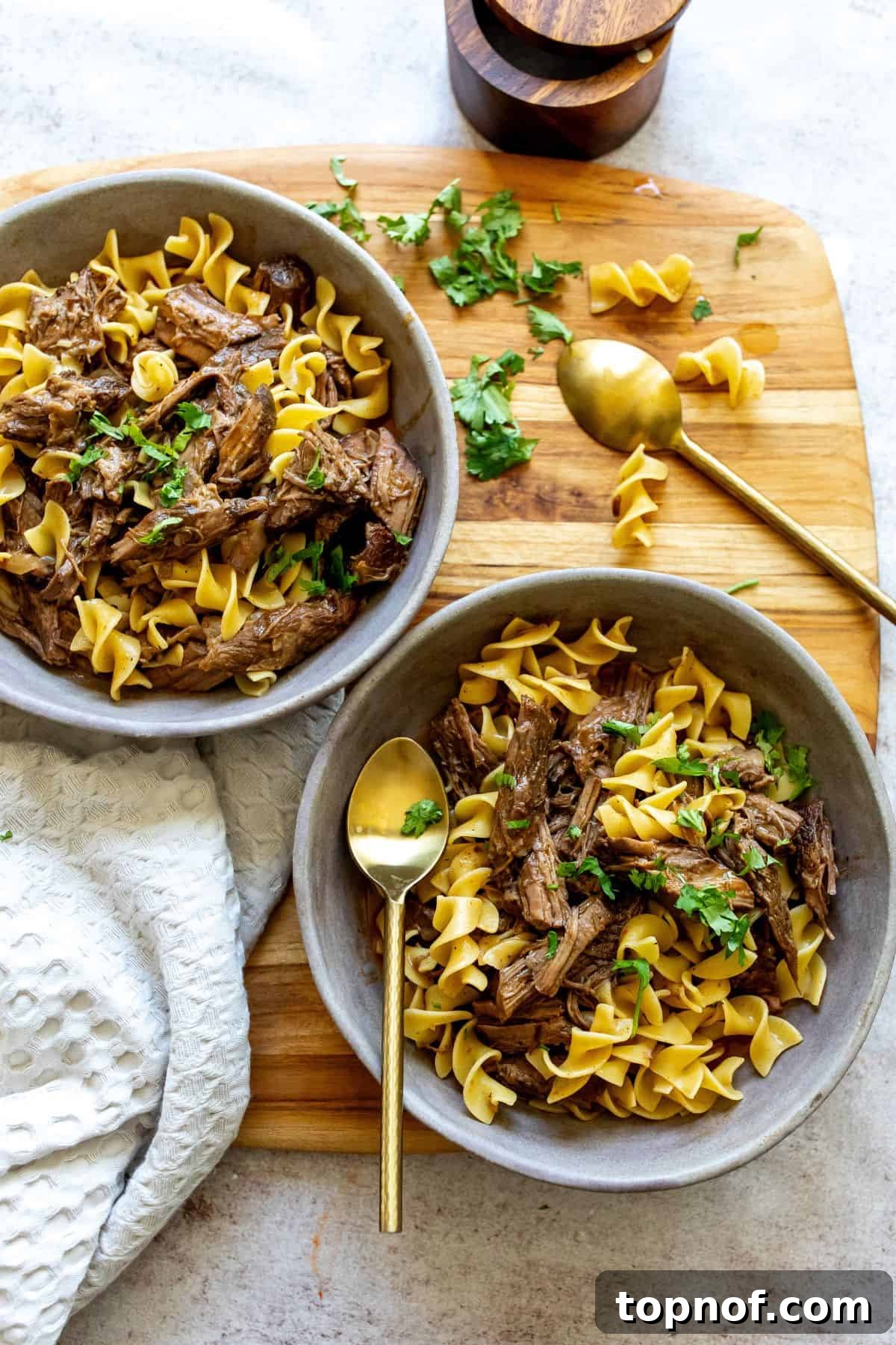 Two grey bowls filled with tender slow cooker beef and noodles, garnished with fresh parsley. A gold spoon rests beside the bowls, inviting you to dig in.