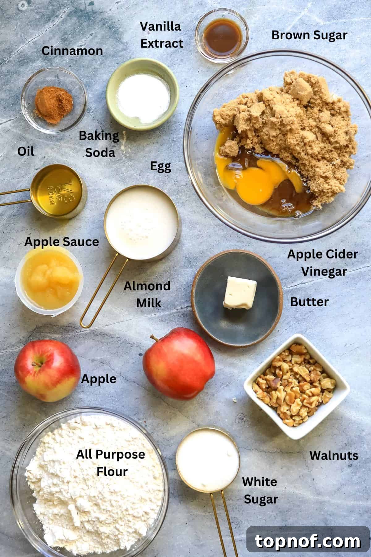 Key ingredients for baking apple cinnamon cake laid out on a kitchen counter, including fresh apples, flour, eggs, and various spices in clear bowls.
