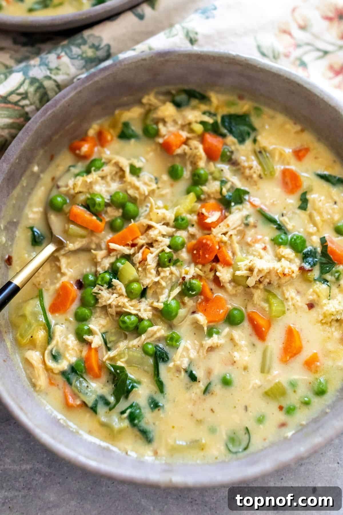 Velvety Chicken & Garden Vegetable Soup 5 Close-up of creamy chicken and vegetable soup in a grey ceramic bowl, showing tender chicken pieces, vibrant vegetables, and a rich broth. A gold spoon is partially submerged.