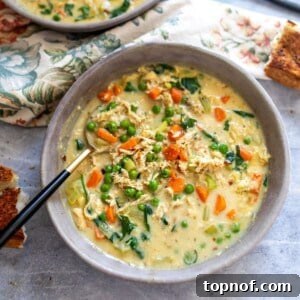 A close-up of a grey bowl filled with creamy chicken veggie soup, rich with tender chicken and colorful vegetables, with a gold and black spoon resting in the bowl.