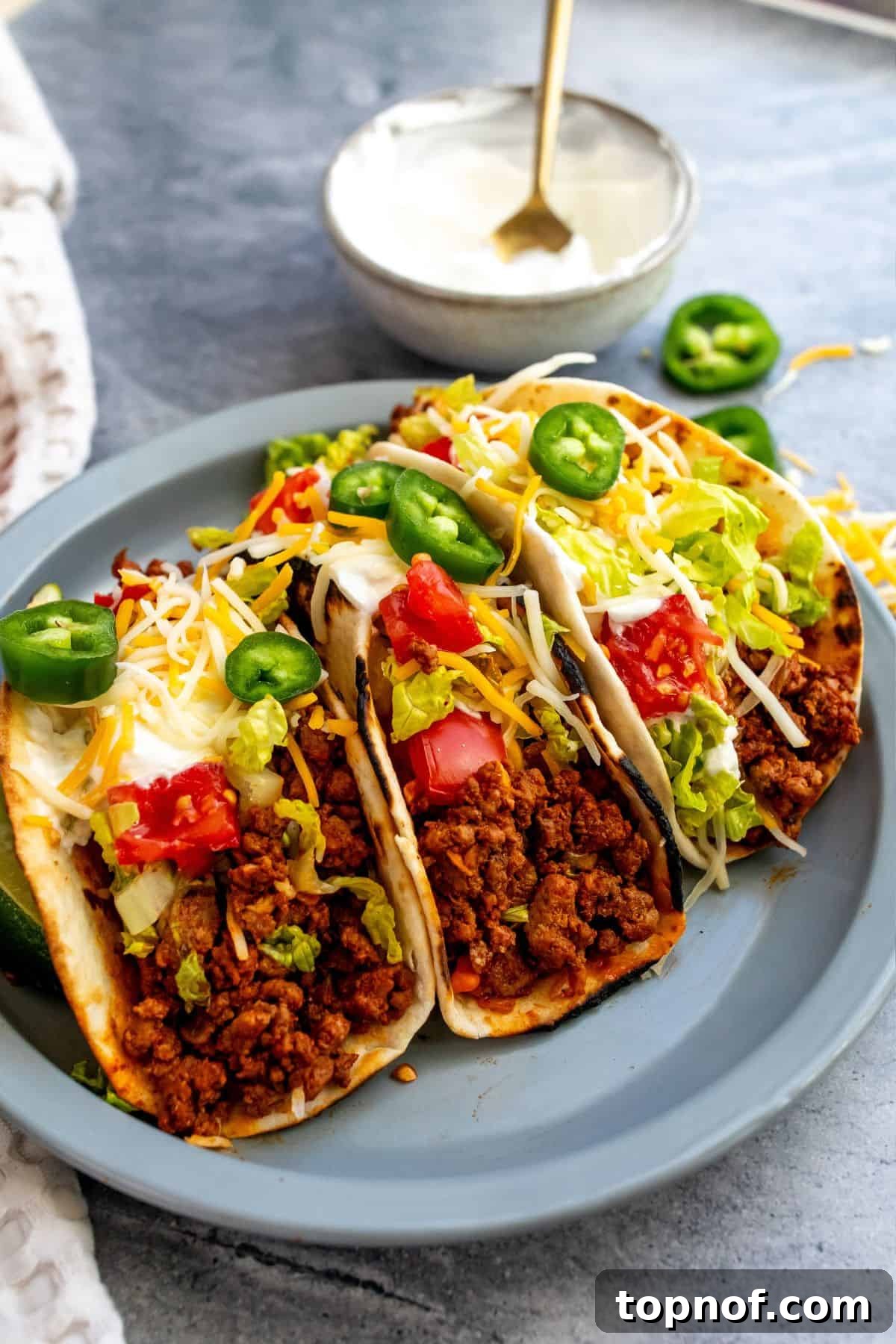 Flavorful Ground Beef Soft Tacos 2 Grey plate topped with three beef tacos in soft tortillas and bowls of cheese, jalapeño and sour cream on the side.