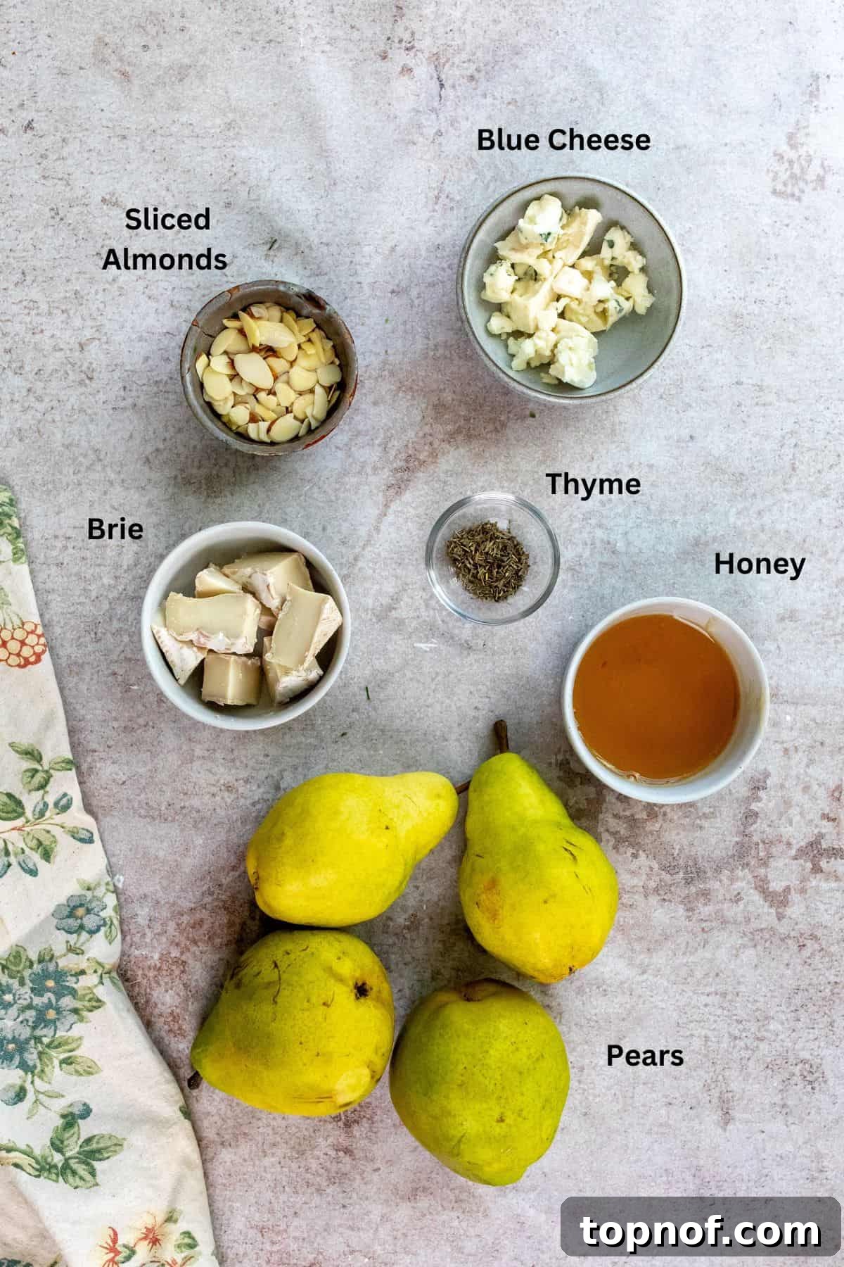 Sweet and Savory Brie Baked Pears 3 A vibrant display of fresh ingredients needed to make baked pears, including whole pears, a wedge of creamy brie, blue cheese crumbles, sliced almonds, and a jar of golden honey, neatly arranged in small mixing bowls on a clean kitchen counter.