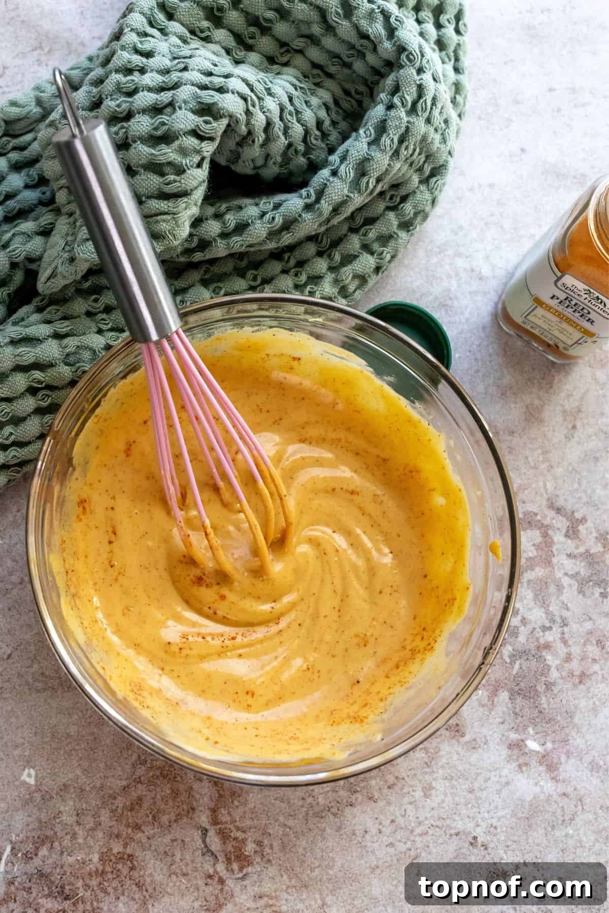 Hot Honey Mustard Sauce 4 A glass bowl with honey mustard and spices being whisked together with a pink whisk, showing the smooth, creamy consistency of the sauce.