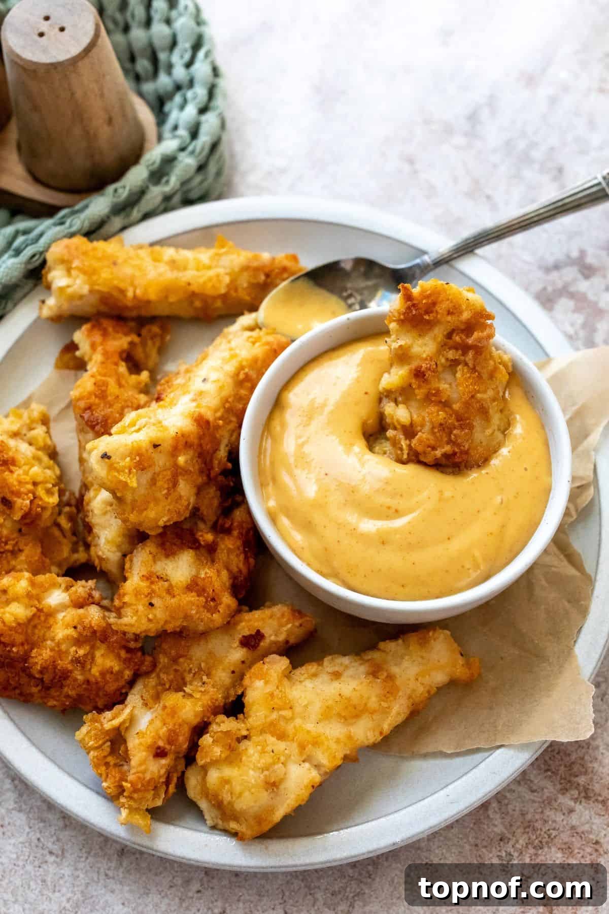 Hot Honey Mustard Sauce 5 Crispy chicken tenders arranged around a white bowl filled with spicy honey mustard, with a silver spoon resting on the side, ready for dipping.