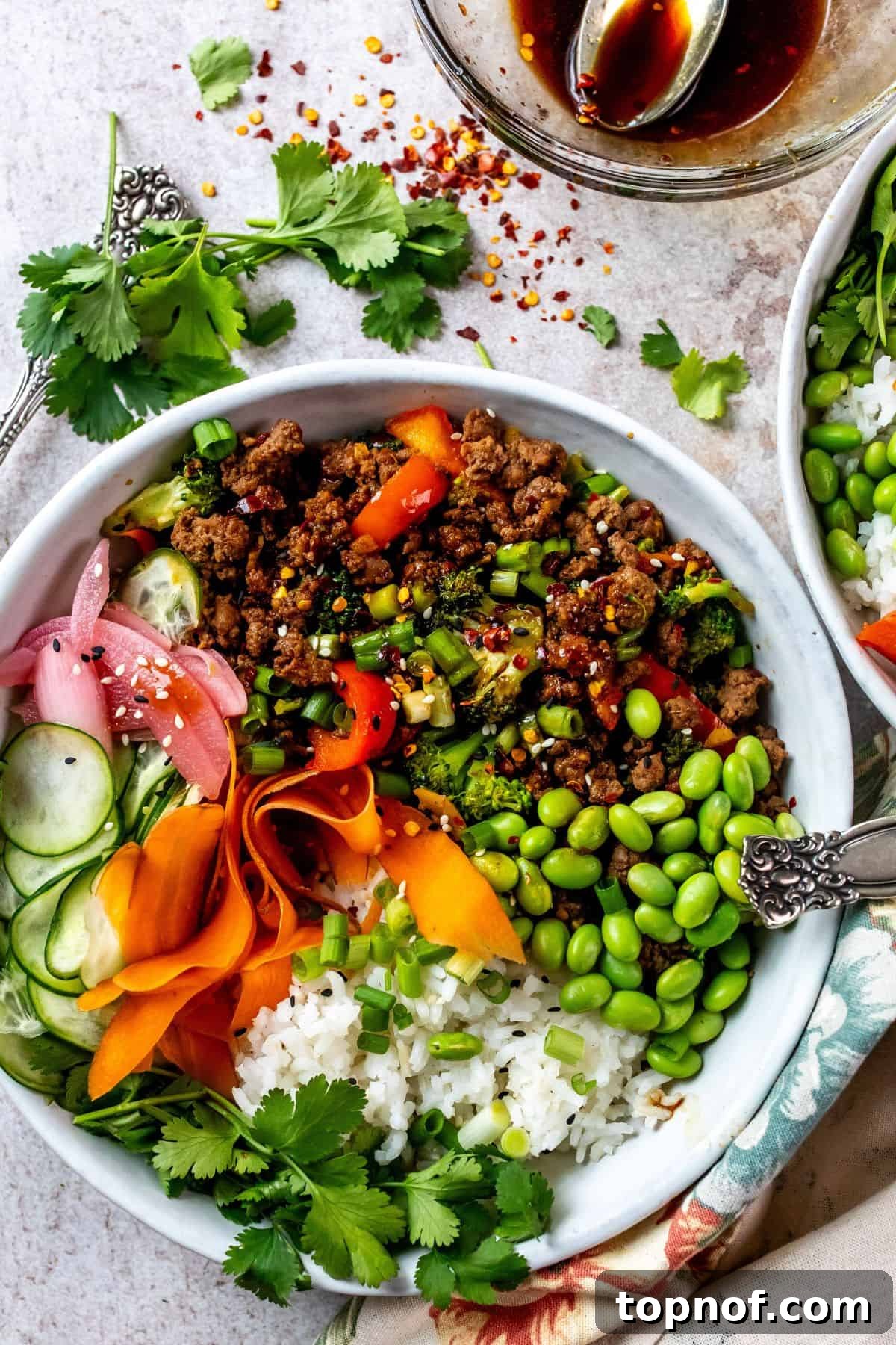 Ultimate Teriyaki Beef Bowls 2 Two white ceramic bowls brimming with succulent ground beef coated in a glossy teriyaki sauce, vibrant fresh chopped vegetables like carrots, cucumbers, and edamame, and a sprinkle of sesame seeds and green onions. A silver fork rests on the side, inviting you to dive in.