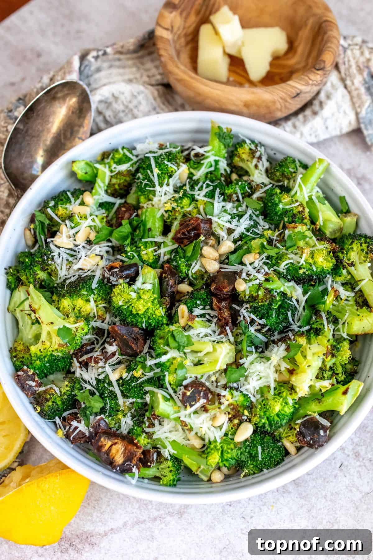 A white bowl filled with charred broccoli, adorned with shredded cheese, lemon zest, chopped dates, and pine nuts, ready to be enjoyed.