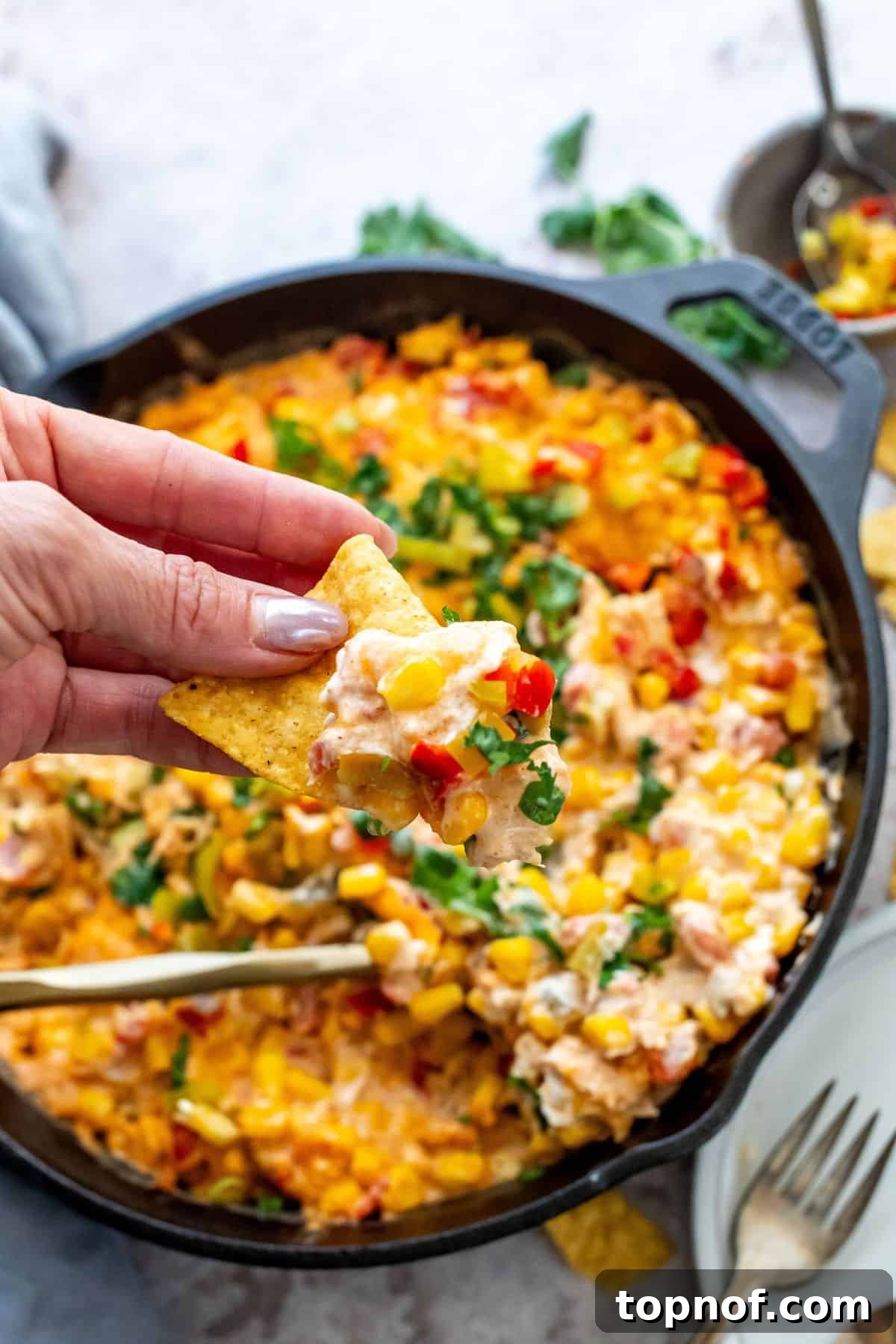 Golden Creamy Corn Delight 5 Black cast iron with corn and cream cheese mixed and topped with cilantro. Gold serving spoon in the mix to serve and a chip.