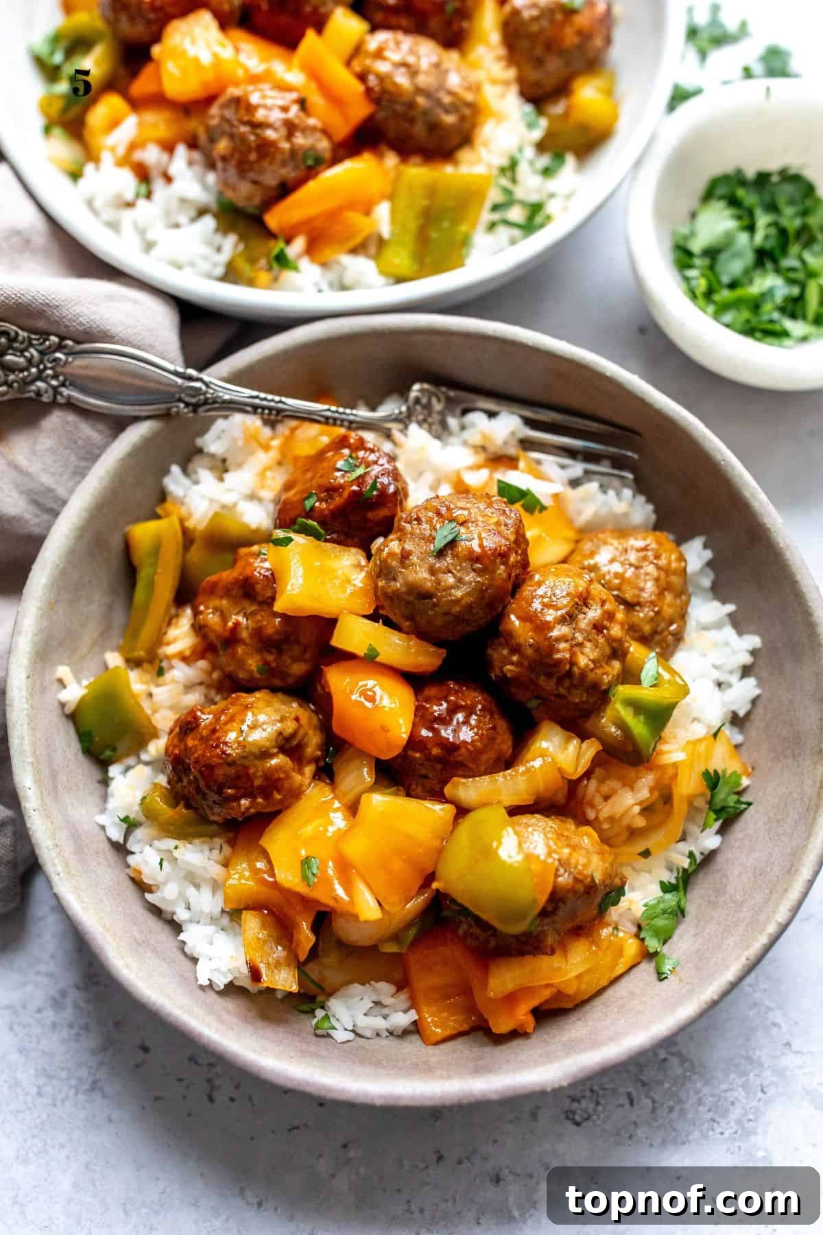Zesty Meatball Bites 2 A grey bowl filled with homemade sweet and sour meatballs and rice, garnished with fresh herbs and a fork resting beside it.