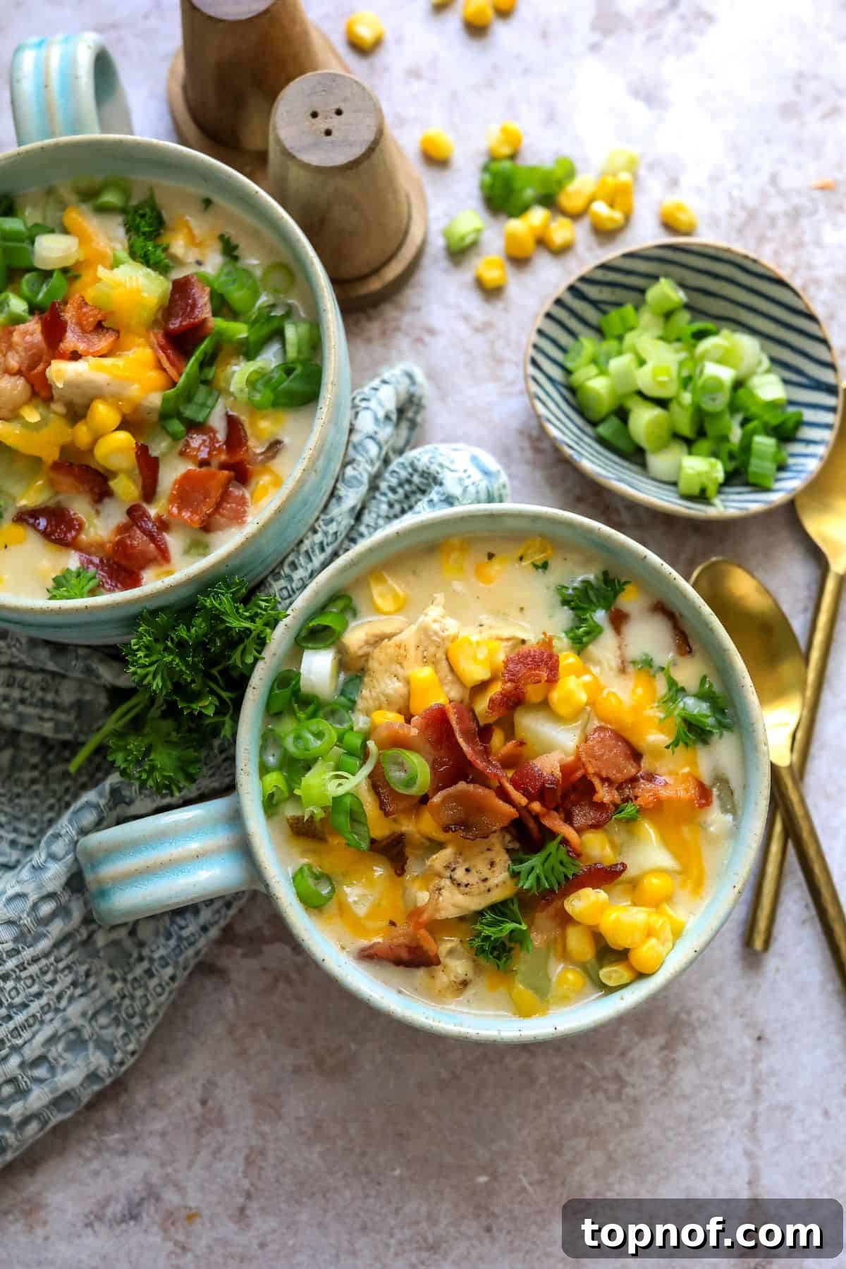Golden Chicken Corn Chowder 2 Two soup mugs with a handle on a counter with corn chowder and bacon on top. Gold spoon to the side.
