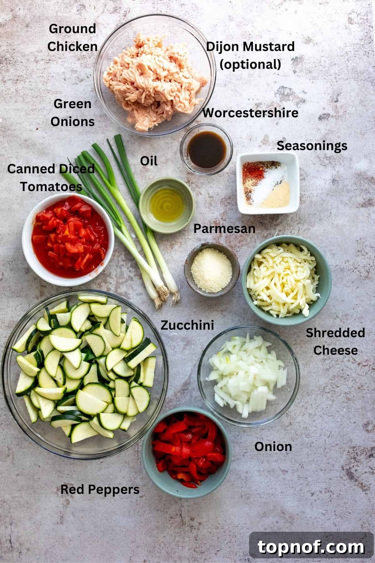 Keto Friendly Zucchini Bake 3 A collection of fresh ingredients laid out on a kitchen counter, including sliced zucchini, diced onions, red bell peppers, ground chicken, canned tomatoes, and various spices, all ready for the Zucchini Bake.