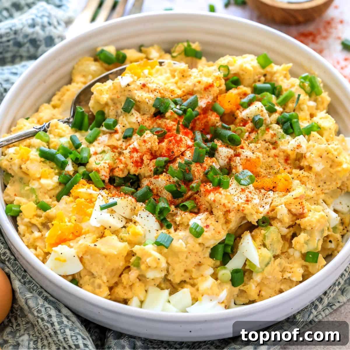 A white ceramic bowl filled with creamy Deviled Egg Potato Salad, garnished with fresh green onions and served with a spoon.