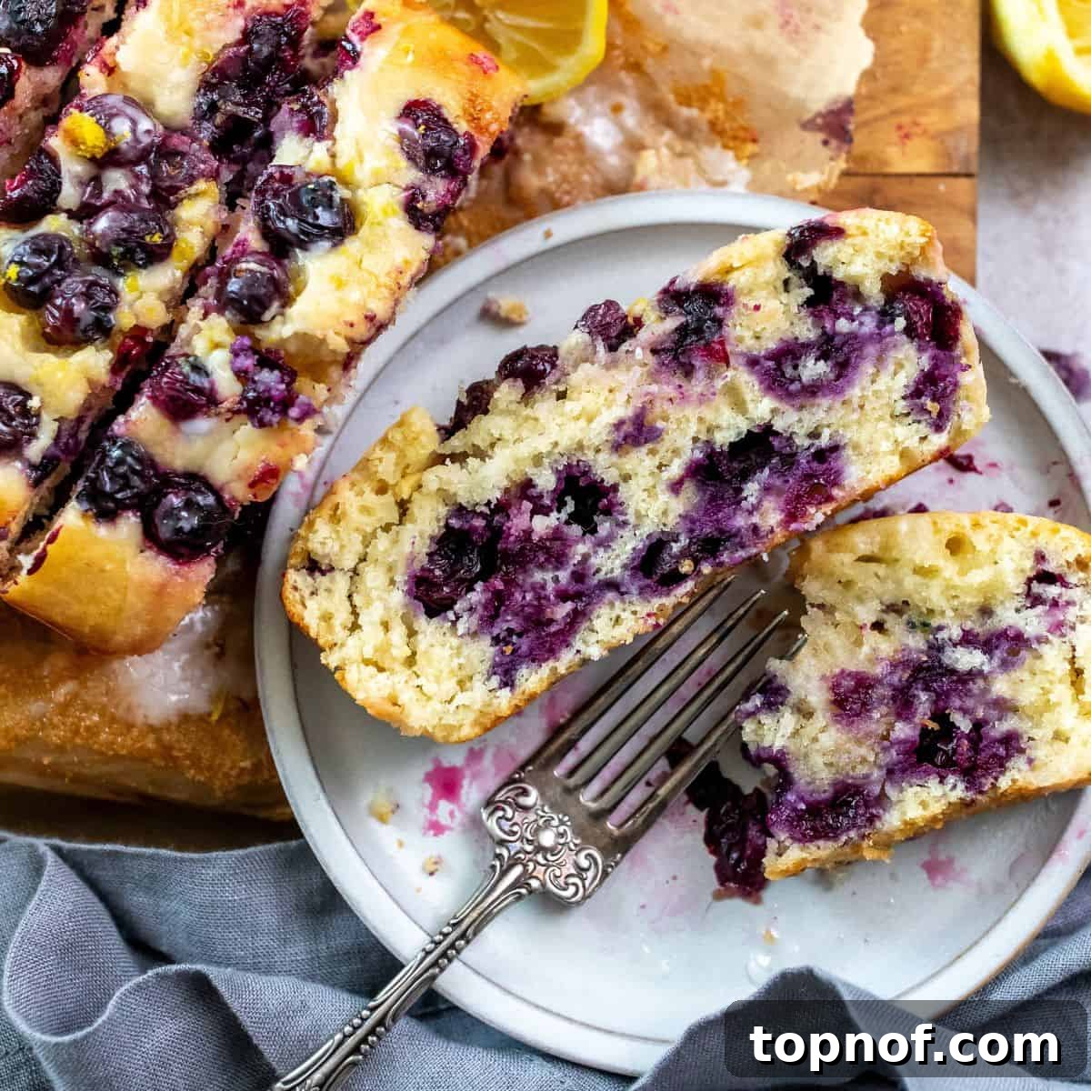 A slice of moist Lemon Blueberry Bread on a plate with a silver fork, accompanied by fresh lemons on the side.