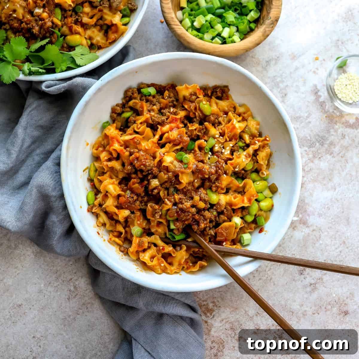 White ceramic bowl filled with saucy spicy garlic noodles and ground beef, garnished with fresh green onions and chopsticks on the side.