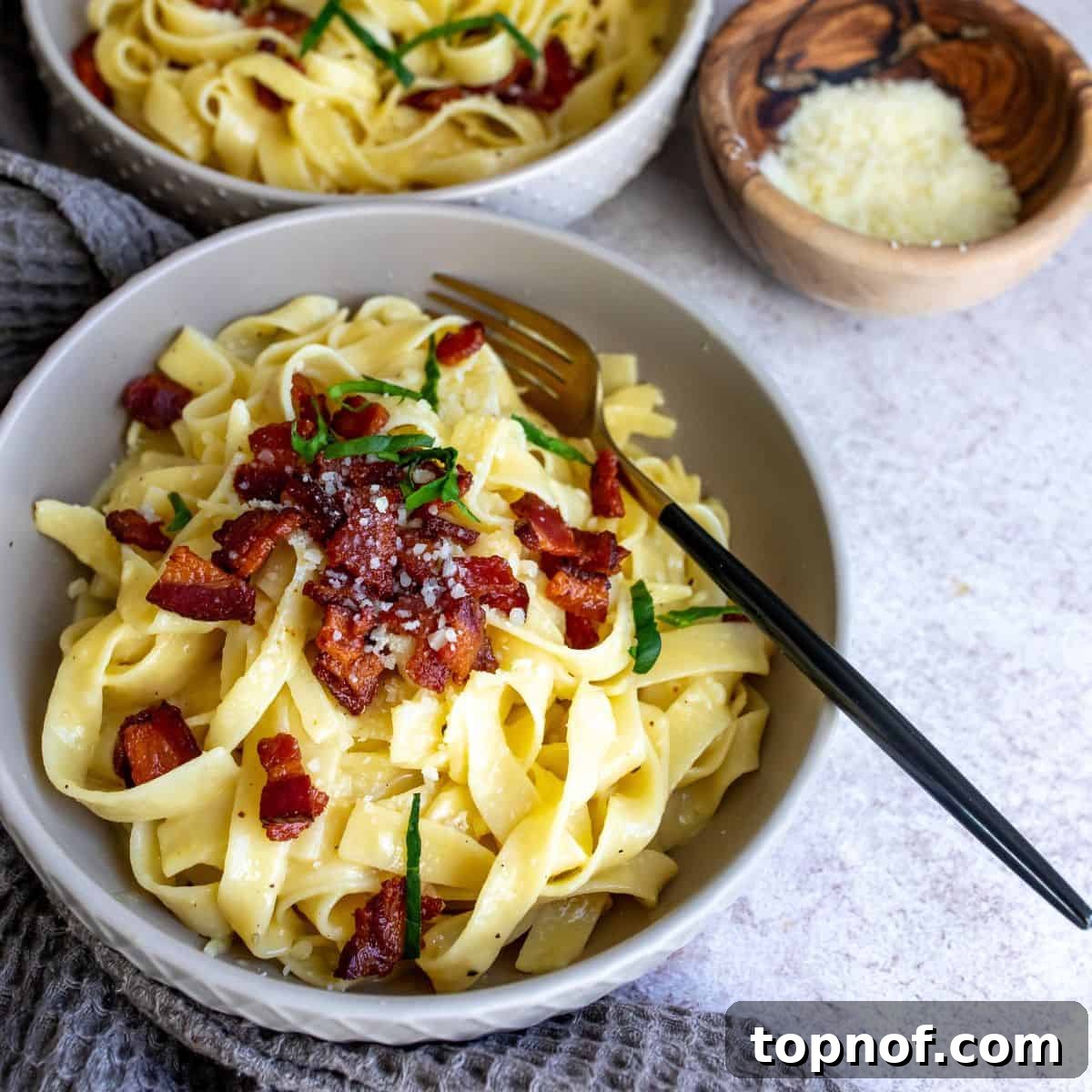 A grey bowl filled with rich Tagliatelle Carbonara, topped with crispy bacon and a fork ready to twirl.