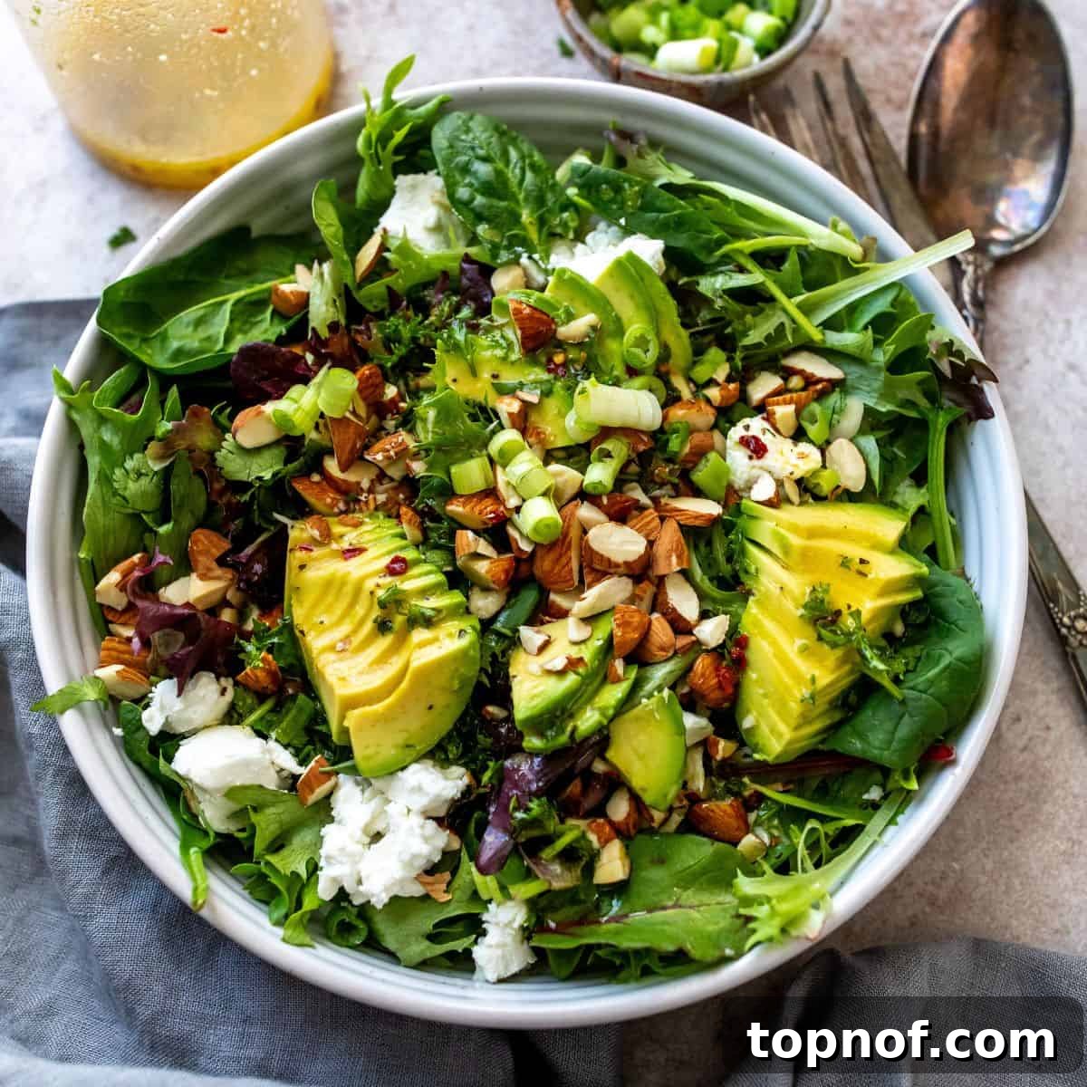 White bowl with mixed greens and avocado on top, with mixing utensils to the side.