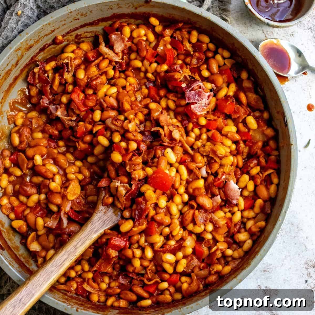 An overhead image of baked beans in a pot with a wooden spoon in the middle, looking rich and savory.