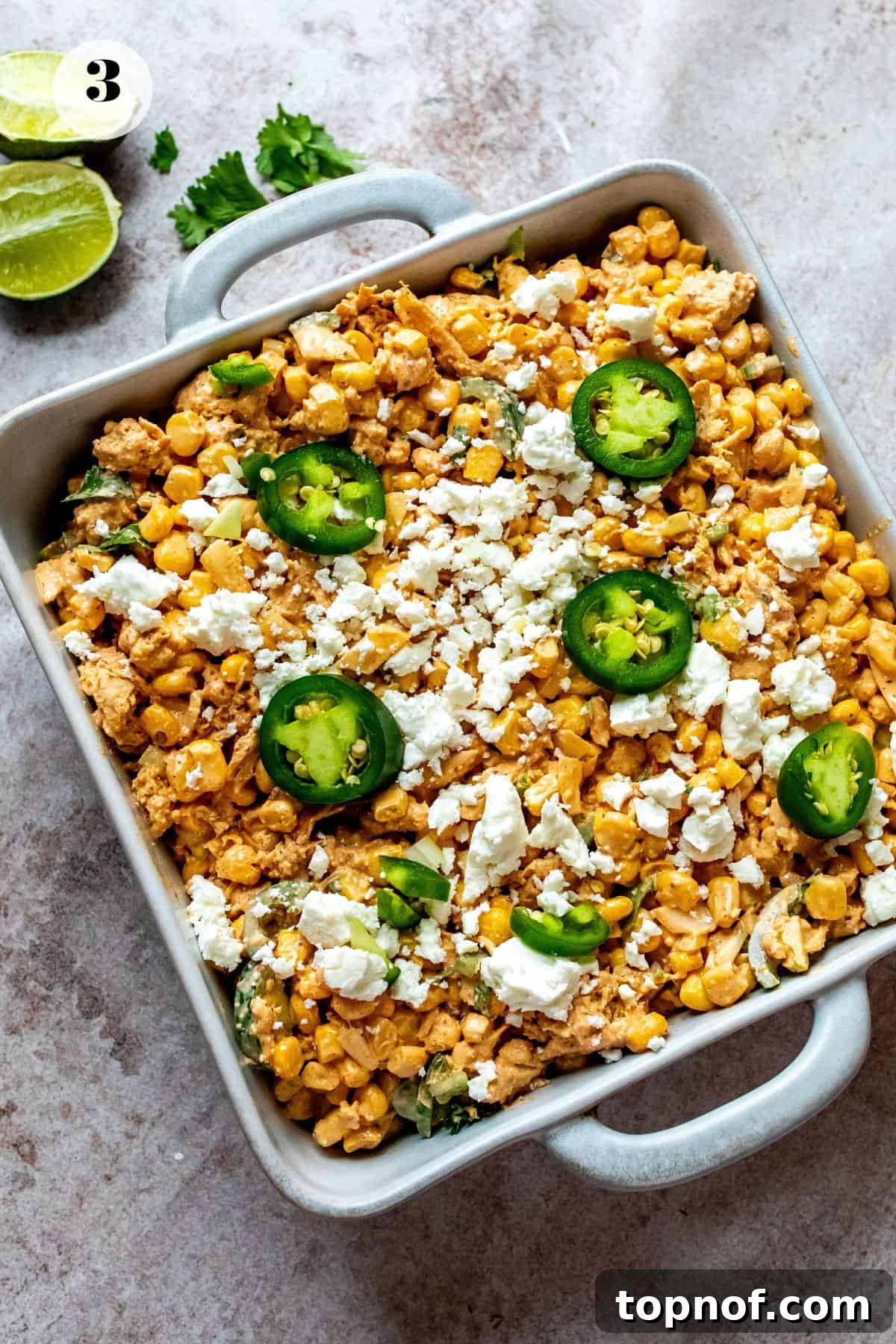 Creamy Elote Bake 5 A close-up view of the Mexican street corn casserole nestled in a white baking dish before it's placed in the oven, showcasing the vibrant corn kernels, diced vegetables, and creamy sauce topped with crumbled cotija cheese and a light sprinkle of chili powder, promising a delicious baked dish.