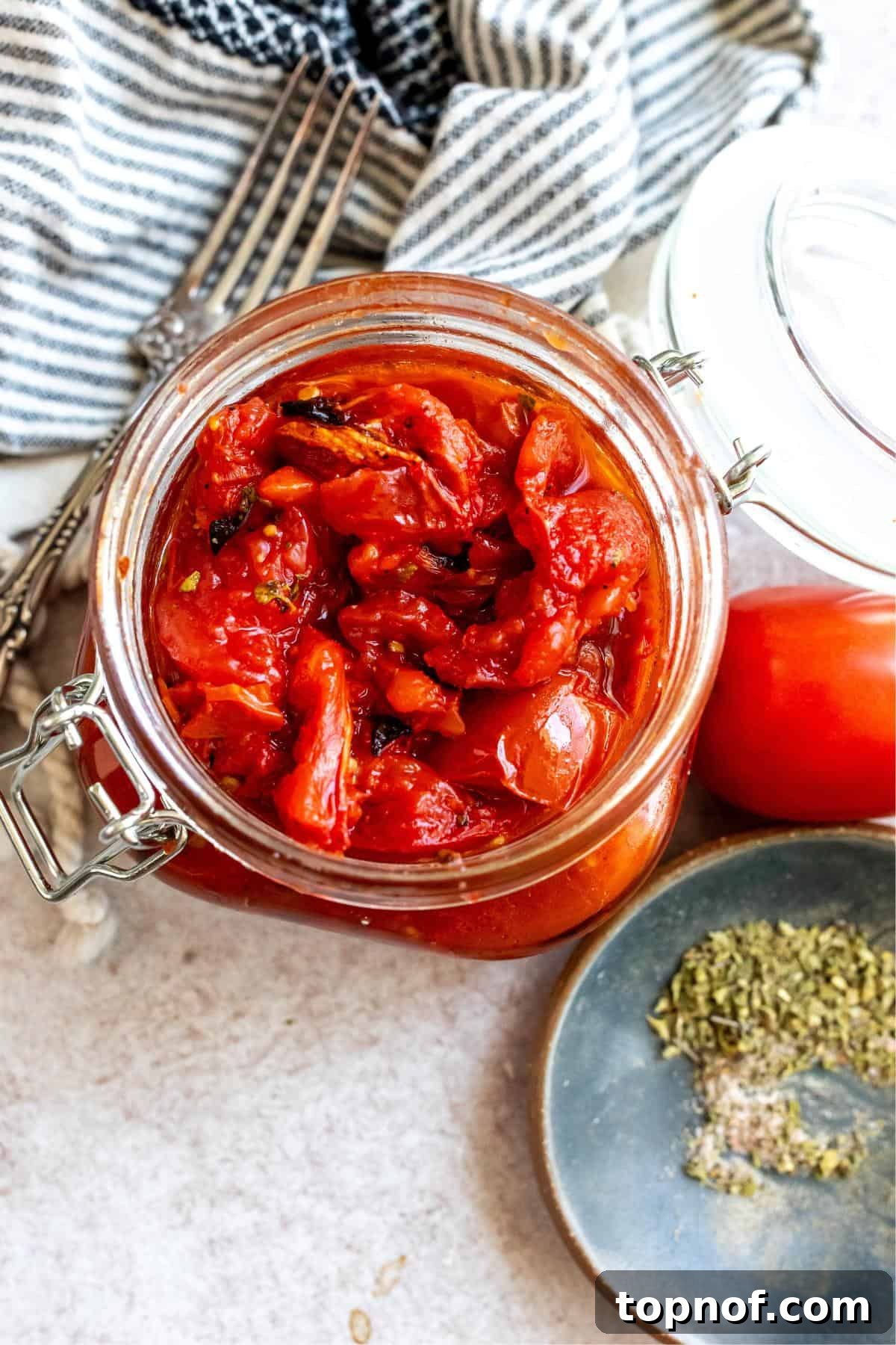 A large glass jar filled with vibrant red homemade fire roasted tomatoes. A fork and dried herbs are artfully placed to the side, highlighting the rustic appeal of the dish.