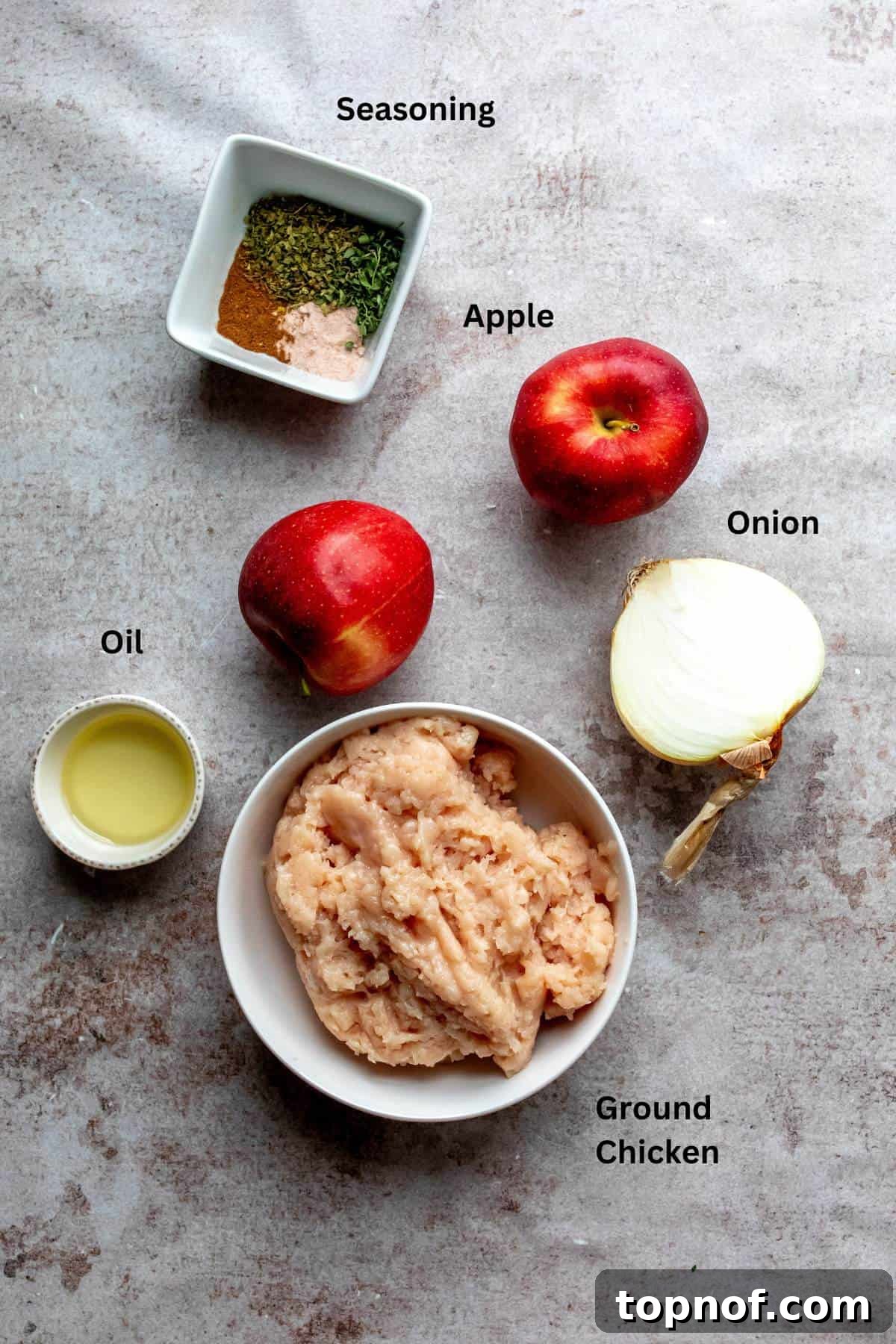 Fresh ingredients laid out on a wooden board: raw ground chicken, vibrant red apples, a white onion, a bottle of olive oil, and various spices in small bowls.