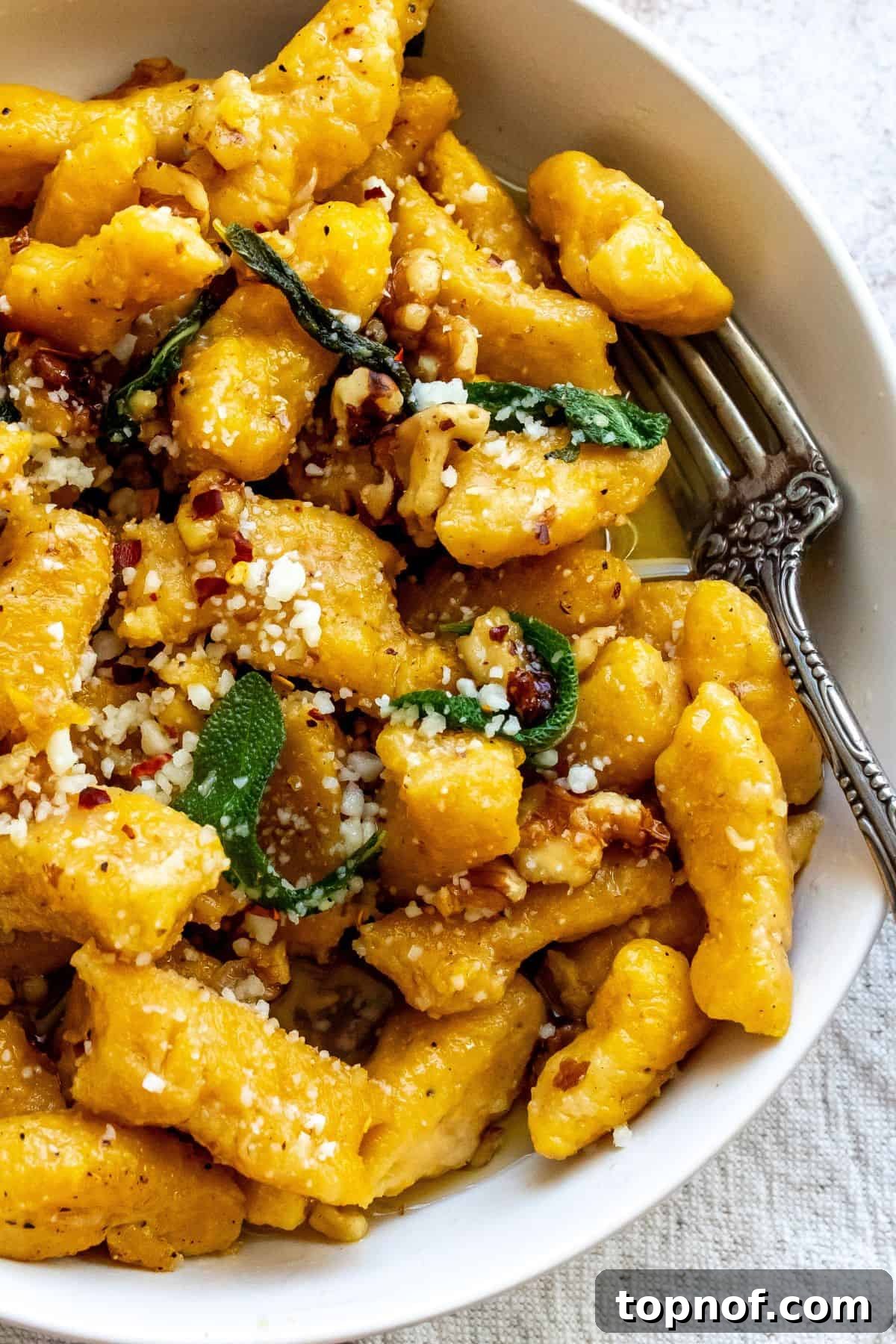 A beautifully plated bowl of butternut squash gnocchi, garnished with fresh sage, walnuts, and Parmesan, with a fork ready to serve.