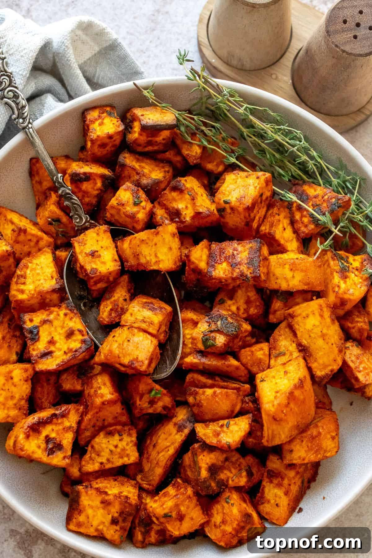 Crispy Baked Sweet Potatoes 2 A bowl filled with beautifully golden and crispy roasted sweet potatoes, garnished with fresh herbs, alongside a rustic silver antique spoon.