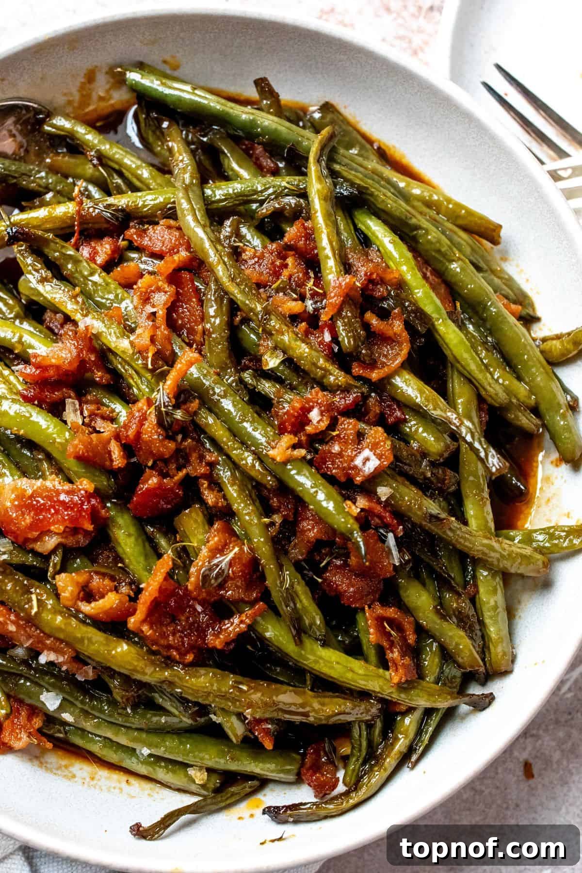 Snappy Green Beans 5 A close-up image of beautifully cooked crack green beans, showing their tender texture and a generous topping of crispy bacon bits, served on a white plate.