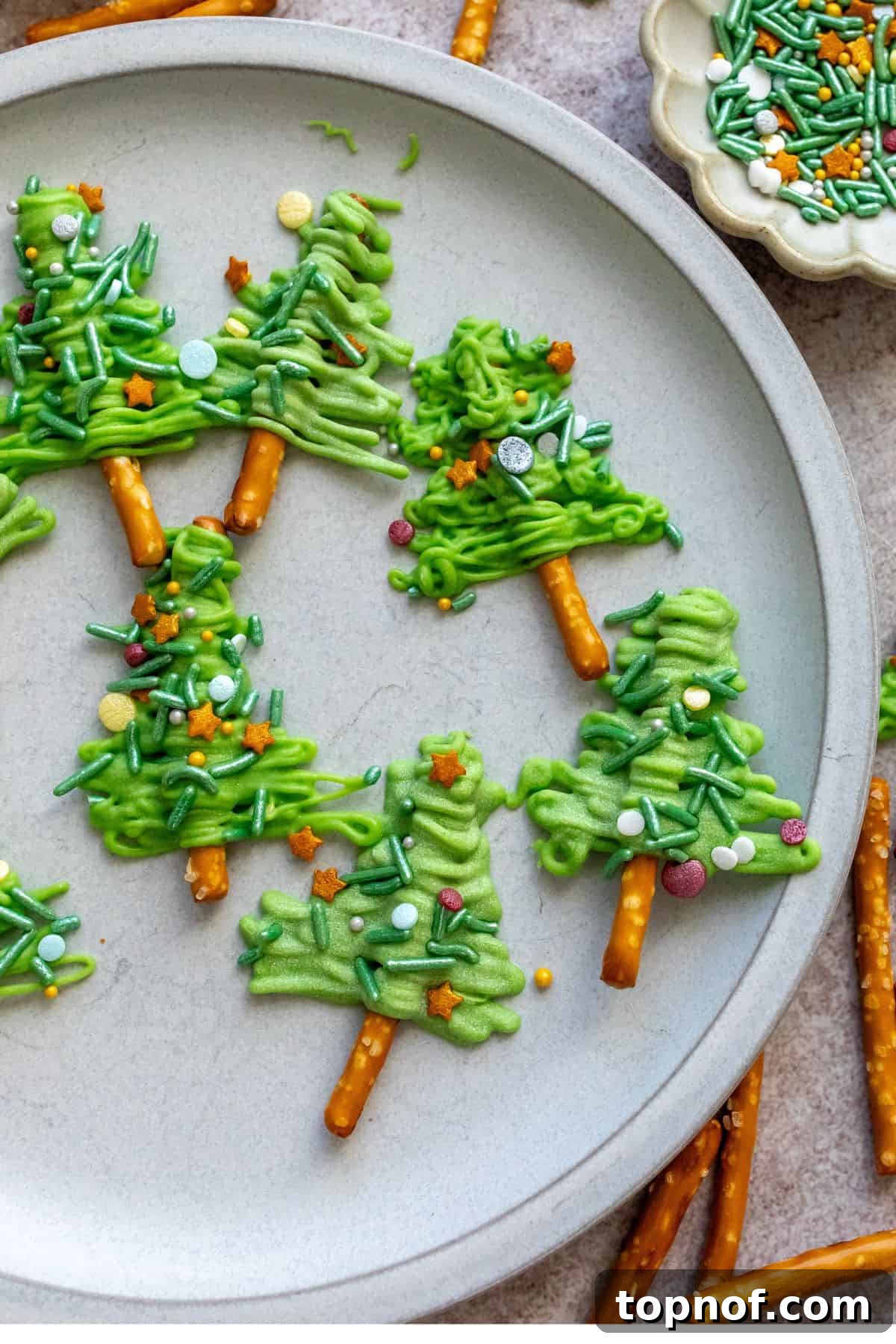 Festive Pretzel Tree Treats 6 Close-up of a white plate with Pretzel Christmas Trees and sprinkles on the side.