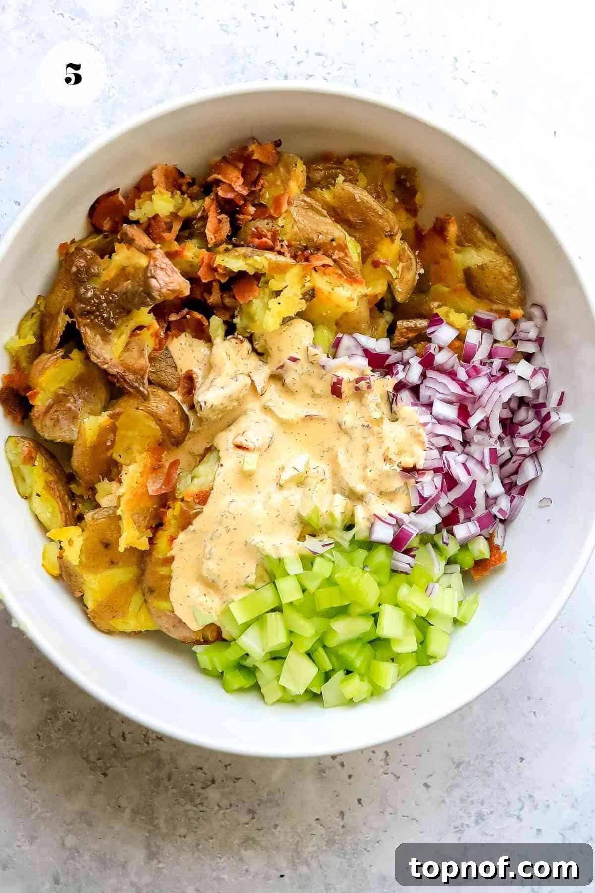 Buffalo Smashed Spud Salad 5 All the fresh ingredients for the Smashed Buffalo Potato Salad, including crispy bacon, red onion, celery, and feta cheese, ready to be mixed in a large white bowl for assembly.