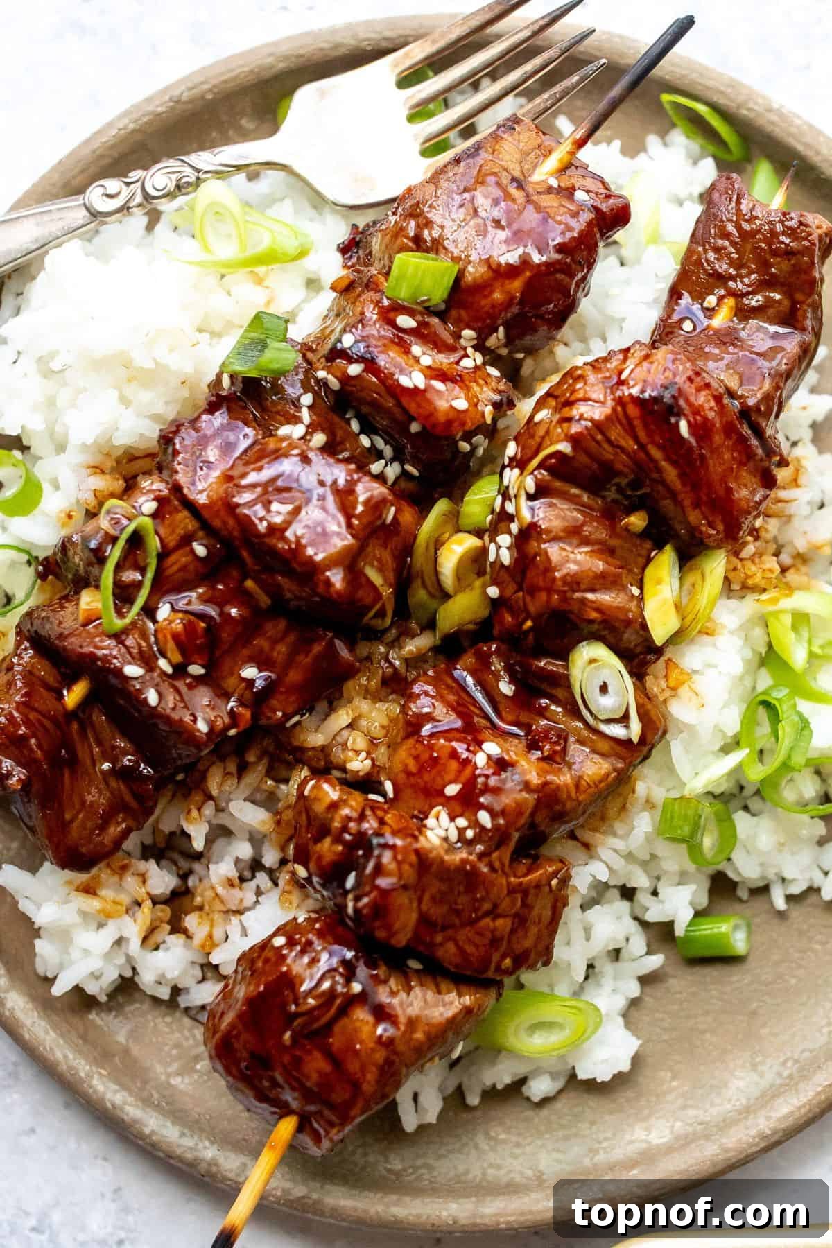 Aromatic Garlic Ginger Beef Skewers 5 A grey plate featuring grilled steak kabobs resting on a bed of fluffy white rice, garnished with fresh green onions.