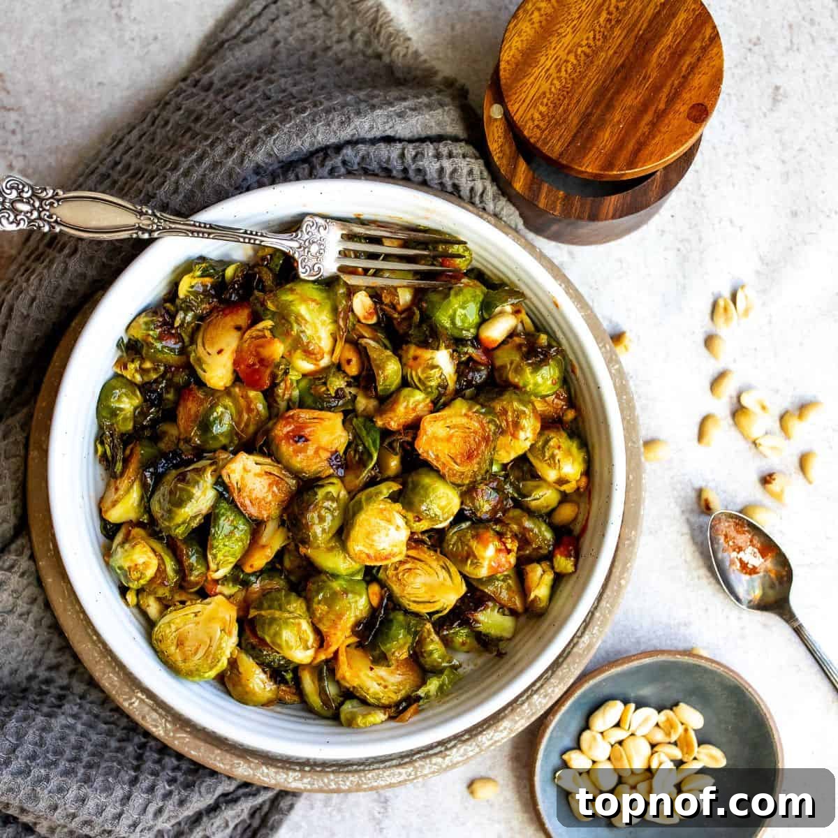 White bowl filled with honey sriracha brussel sprouts and sauce and a silver spoon with sauce. Peanuts on the side. 