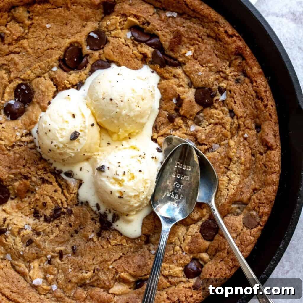 A freshly baked Cast Iron Cookie still in its black skillet, generously topped with two scoops of creamy vegan vanilla ice cream and two spoons, inviting immediate enjoyment and sharing.