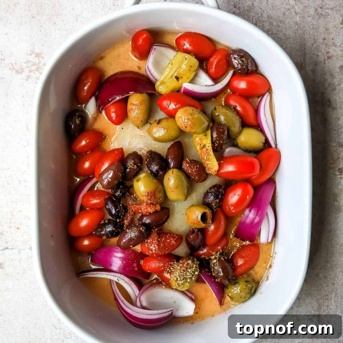 Herbed Mediterranean Fish Bake 4 White fish fillet in a baking dish, topped with a colorful medley of cherry tomatoes, olives, and sliced red onions, ready for baking.