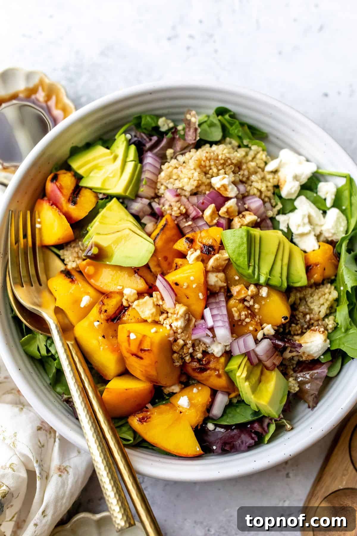 Juicy Peach and Tangy Feta Salad 2 A beautifully arranged white bowl filled with Peach Feta Salad, topped with fresh avocado slices and crumbled feta cheese. Serving utensils rest beside the bowl, inviting you to dig in.