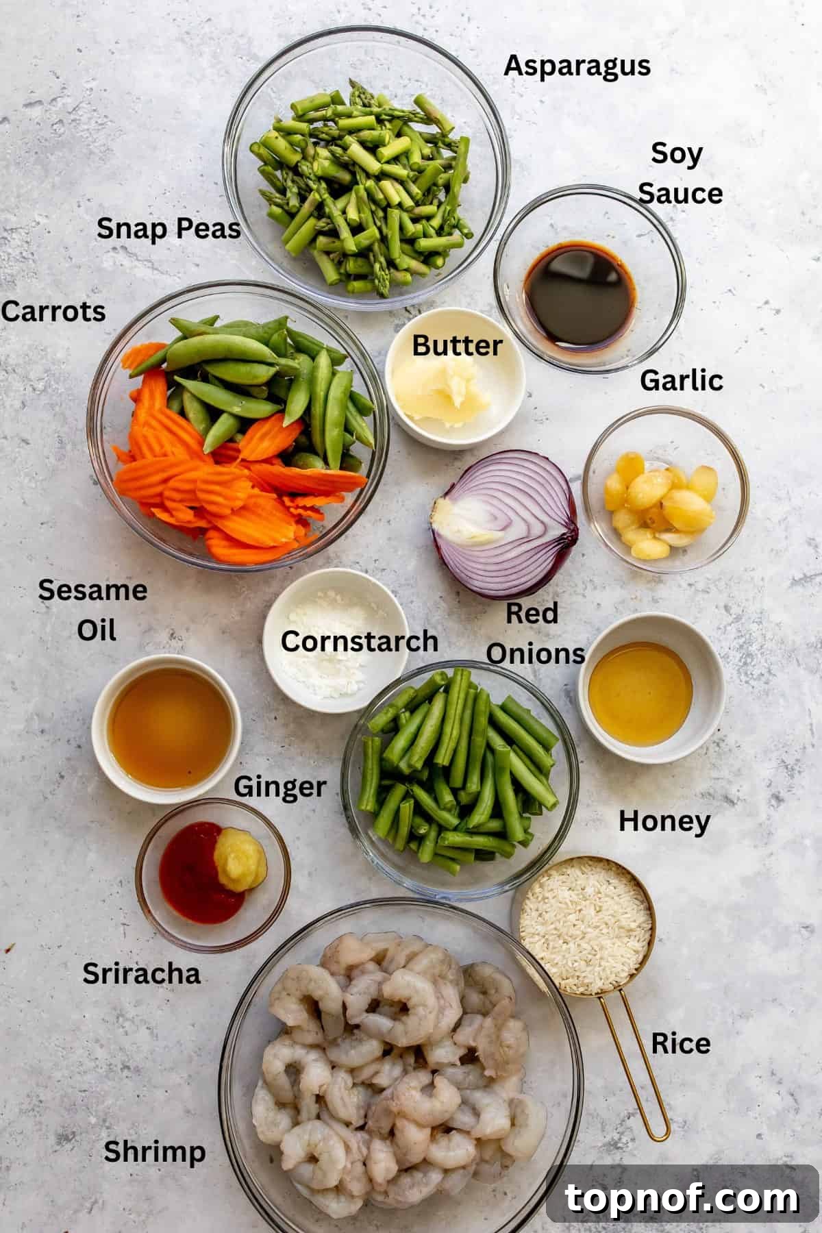 Neatly arranged bowls of fresh ingredients for garlic shrimp stir fry, including raw shrimp, chopped asparagus, carrots, and various sauce components.