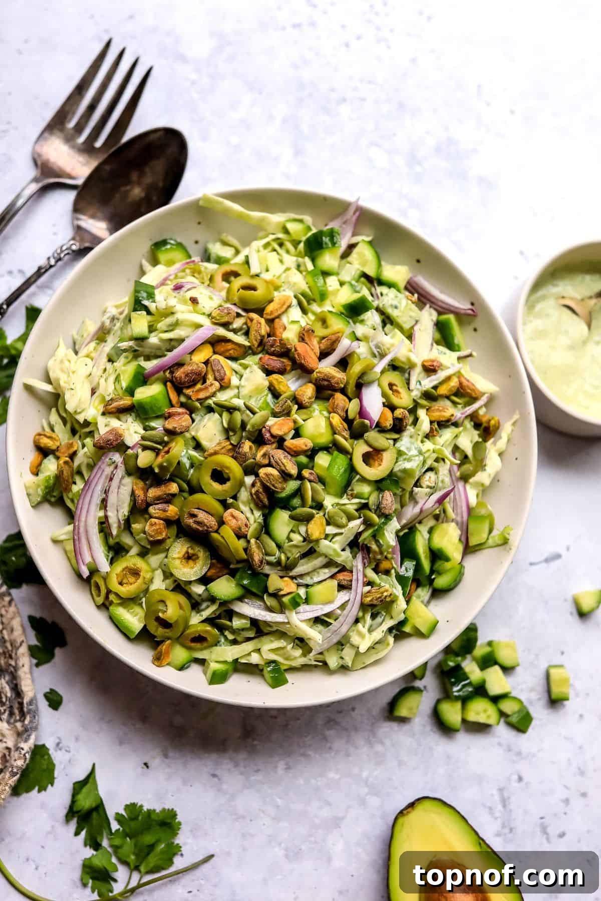 Emerald Goddess Salad 2 A large serving bowl filled with a vibrant Green Goddess Salad, featuring thinly sliced cabbage, cucumbers, celery, red onion, and green olives, with a small bowl of creamy green dressing placed elegantly to the side.
