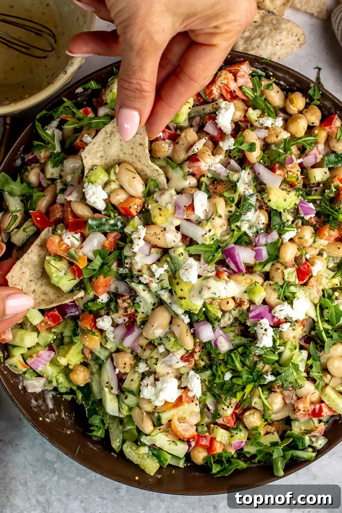 White bean salad in  brown bowl with a hand dipping a tortilla chip into it.